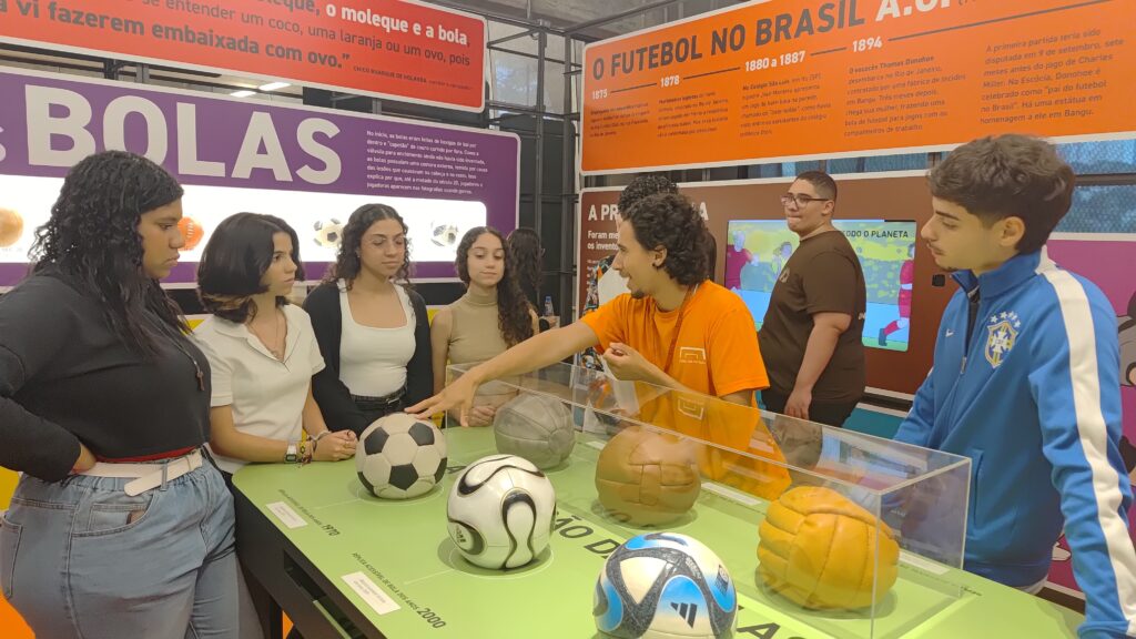 Um grupo de jovens escuta um educador do Museu apresentando uma cole&ccedil;&atilde;o de bolas de futebol colocadas em uma mesa verde, que fica no meio da exposi&ccedil;&atilde;o principal, em um ambiente muito colorido.S&atilde;o mo&ccedil;as e rapazes de perfis variados. O educado toca uma das bolas com a m&atilde;o.